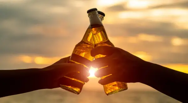 Dwie ręce wznoszą butelki piwa do toastu o zachodzie słońca. W Niemczech od ilu lat można pić alkohol? Od 16 roku życia.