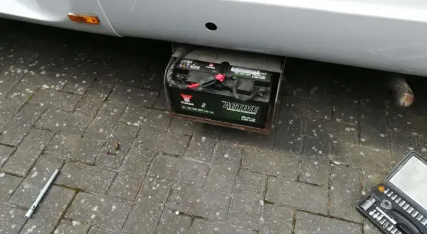 Emplacement batterie Ford Transit : une batterie Yuasa "Active Leisure & Marine" est visible sous le véhicule, dans un support métallique. Une clé à douille et une boîte à outils sont au sol.