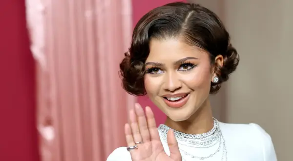 Uśmiechnięta Zendaya w białej sukni i biżuterii, macha ręką. Fani zastanawiają się, czy Zendaya jest w ciąży.