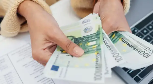 Mani che contano banconote da 100 euro, forse per la divisione conto corrente eredi. Documenti e laptop sullo sfondo.