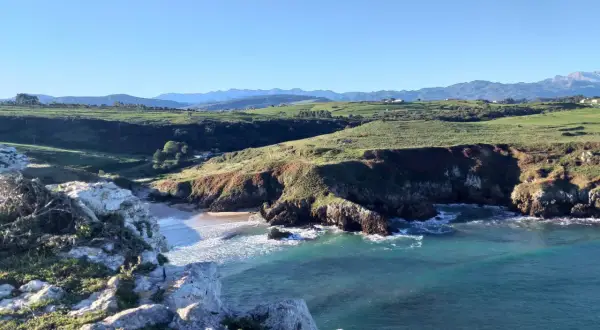 Acantilados y mar turquesa, ideales para rutas senderismo San Vicente de la Barquera. Paisaje costero con verdes colinas y montañas al fondo.