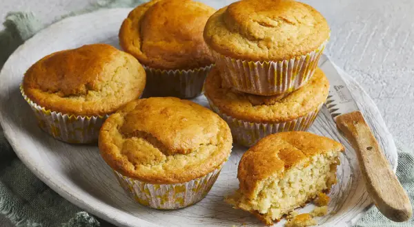 Goldbraune Buttermilch Muffins auf einem Holzteller, einer davon angebrochen, mit einem Messer daneben.