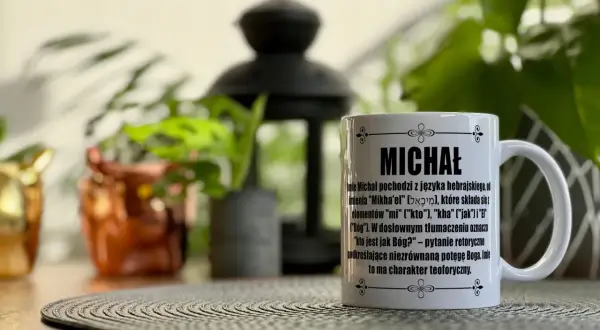 Biały kubek z napisem "MICHAŁ" i opisem znaczenia imienia, idealny na imieniny Michała. W tle rośliny i ozdobna latarenka.