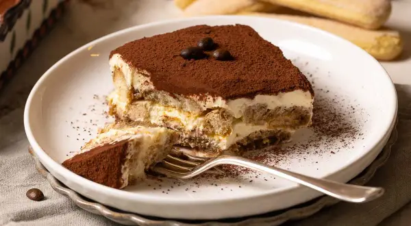 Ein Stück Tiramisu, zubereitet nach einem einfachen tiramisu rezept, liegt auf einem Teller. Kakaopulver bestäubt die cremige Schicht und die Löffelbiskuits.