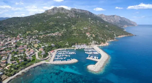 Baunei: un porto turistico con barche ormeggiate, case colorate e montagne verdi che si tuffano nel mare cristallino. Le spiagge vicine invitano al relax.