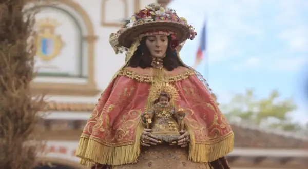 Virgen del Rocío pastora, con su manto rosa bordado y el Niño Jesús en brazos, adornada con flores y un sombrero.
