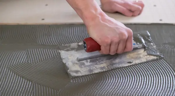 A hand uses a notched trowel to spread thinset for cement board, creating a textured surface for tile adhesion.