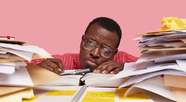 Un homme fatigué, entouré de piles de documents, semble avoir un avis de départ. Il repose sa tête sur un livre ouvert, l'air dépassé par la paperasse.