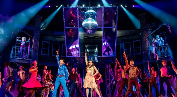 Tancerze w disco strojach na scenie, oświetleni kolorowymi światłami. W tle lustrzana kula i obrazy z tańczącymi postaciami, nawiązujące do gorączka sobotniej nocy utwory.