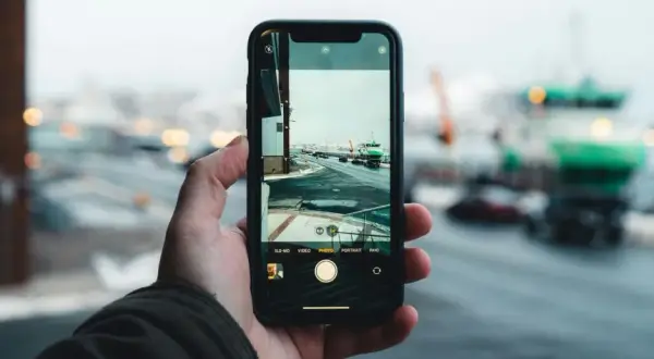 Dłoń trzyma smartfon, na ekranie którego widać interfejs aparatu. Pokazuje, jak dziala aparat, rejestrując zimowy krajobraz z drogą i zielonym statkiem.