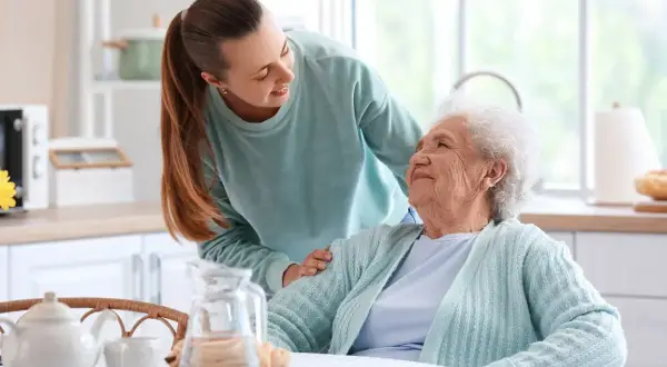 Młoda kobieta wspiera starszą panią przy stole. Rozmowa o tym, ile wynosi zasiłek opiekuńczy, jest ważna dla zapewnienia godnego życia seniorom.