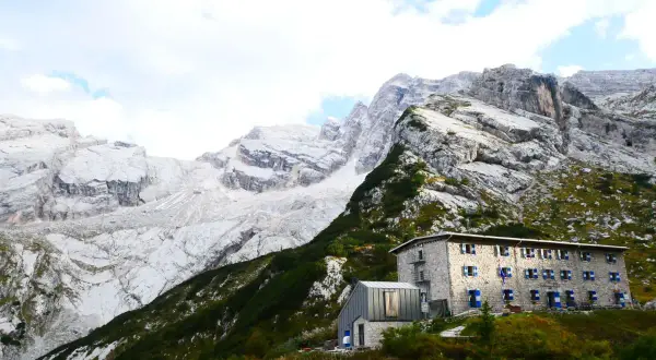 Un rifugio in pietra con persiane blu si staglia su un pendio verdeggiante, sotto imponenti montagne rocciose. Un esempio di rifugi in Veneto.