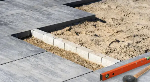 Układanie płyt betonowych na piasku, z widocznym krawężnikiem i narzędziami budowlanymi.