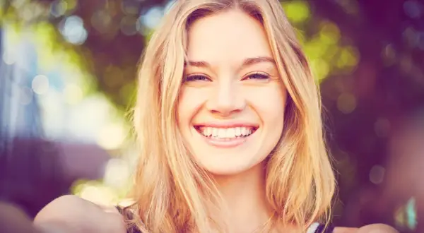 Strahlendes Lächeln einer jungen Frau mit blonden Haaren, die eine natürliche Frisur trägt.