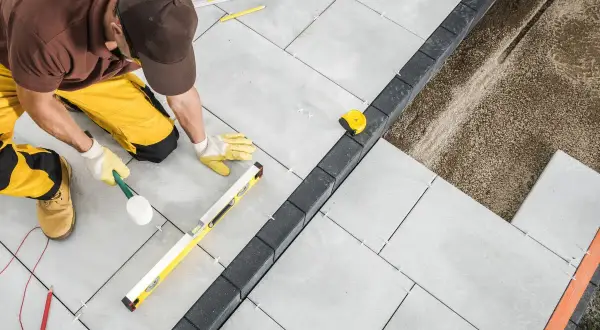 Operaio con martello di gomma e livella esegue la posa a secco di pavimenti esterni in lastre grigie, creando un bordo con pietre scure.