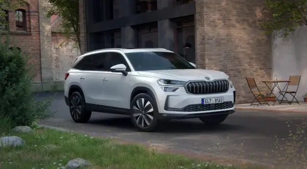 Biały Skoda Kodiaq 2025 zaparkowany przed ceglanym budynkiem, z zielenią i stolikiem z krzesłami w tle.