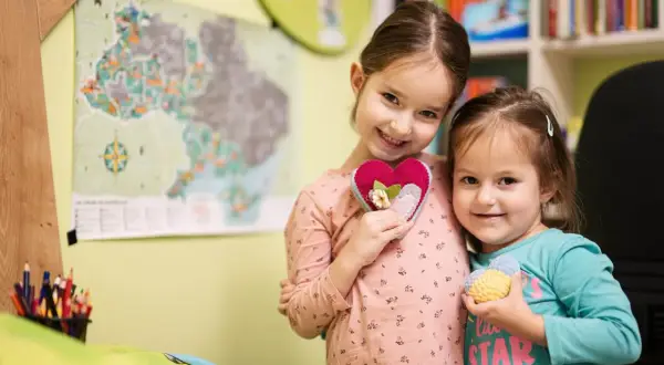 Dwie uśmiechnięte dziewczynki trzymają serduszka. Starsza w różowej bluzce, młodsza w turkusowej. W tle mapa i półka z książkami. Ciekawi ich, ile godzin w zerówce spędzają na zabawie.