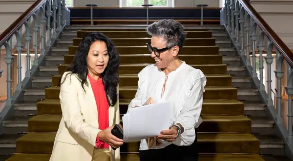 Två kvinnor diskuterar papper på en trappa. Den ena ger den andra en telefon, kanske för att hantera administrativa uppgifter.