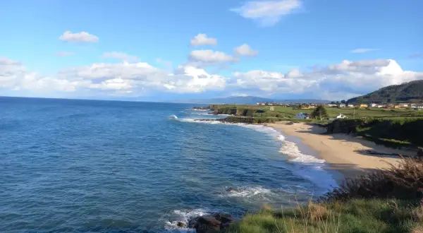 Mar azul y cielo nublado sobre la playa de Xuncos, con casas dispersas en la costa verde.