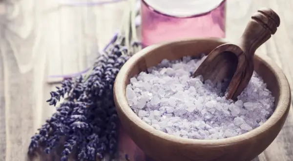 Lavendel und Kristallsalz in einer Holzschale, perfekt zum **Badesalz selber machen**. Ein Glas mit lila Flüssigkeit steht daneben.