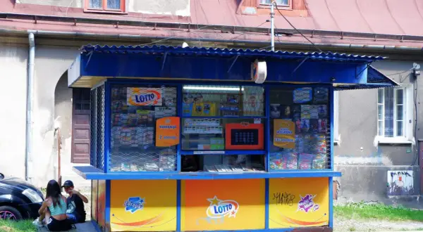 Kiosk Lotto z ekspozycją zdrapek i informacją o kumulacji. Dzieci siedzą obok, zastanawiając się, dlaczego kolektorzy nie chcą sprzedawać zdrapek z konkretnych numerów paczek.