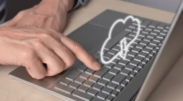 Dłoń naciska klawisz na laptopie, nad którym unosi się symbol chmury internetowej z ikoną pobierania.