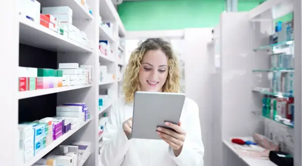 Farmacéutica sonriente revisa inventario en su tablet, rodeada de estantes llenos de productos de atida mifarma.