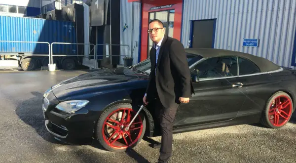 Man i kostym med glasögon vid en svart cabriolet med röda fälgar. Han håller i en fälgnyckel, redo att dra åt efterdra hjulbultar.
