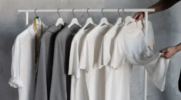 En vit skjorta, grå hoodie och flera vita t-shirts hänger på en klädställning. En hand plockar ner en av t-shirtarna, som en del av en basgarderob herr.