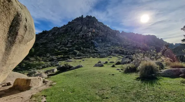 Majestuosa montaña rocosa en la ruta Soto del Real, con un prado verde y el sol brillando en un cielo azul con nubes.