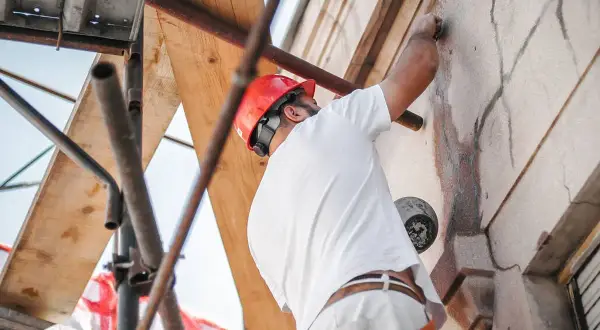 Obrero con casco rojo repara grietas en una fachada desde andamios.