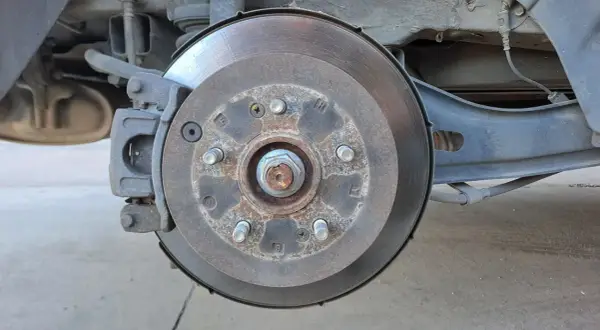 Gros plan sur un disque de frein usé, rouillé et marqué par l'usage. L'étrier de frein est visible, ainsi que des éléments de suspension.
