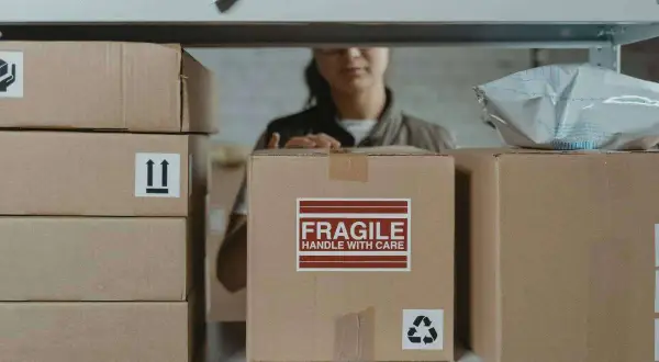 Pakowacz ręczny, kod zawodu 432301, przygotowuje paczki do wysyłki. Na zdjęciu widać stosy kartonów, jeden z napisem "FRAGILE".