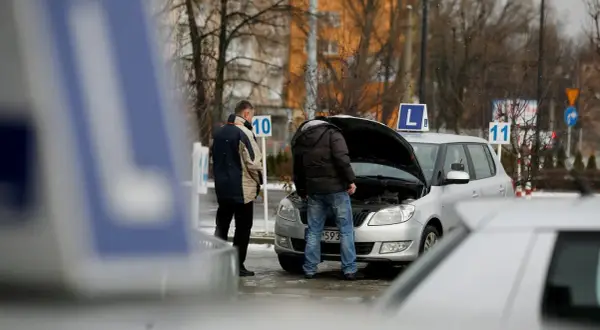 Dwóch mężczyzn przy samochodzie z otwartą maską, znaki "L" i numery na placu manewrowym.