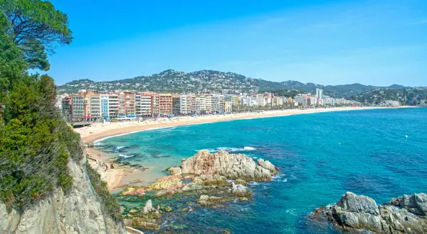 Widok na plażę i zatokę w Lloret de Mar. W oddali widać zabudowania, a na pierwszym planie skaliste wybrzeże i turkusowe morze. Idealne miejsce na wakacje, niedaleko lotniska.