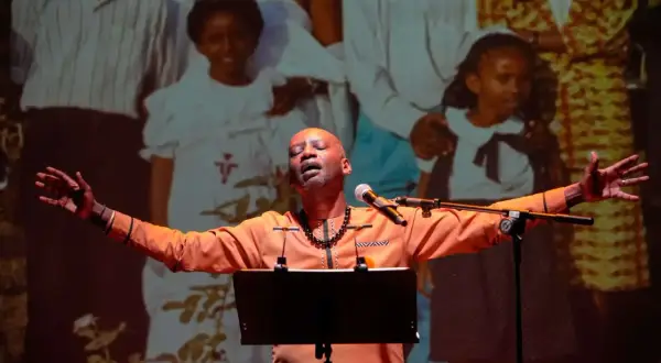 A man in an orange shirt sings with his arms outstretched in front of a projection of a family.