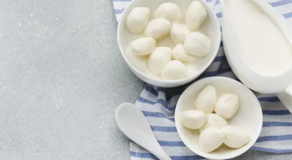 Dwa białe naczynia z serkiem fior di latte, jedno z kulkami w zalewie, obok dzbanek z mlekiem i łyżka na tle z niebiesko-białym pasem.