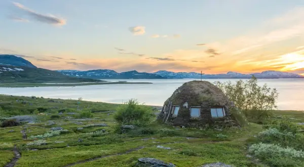 En grästorvstuga med kors på toppen vid en sjö i Padjelanta nationalpark, Sverige, under en solnedgång.