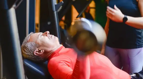 Starszy mężczyzna wykonuje trening funkcjonalny ćwiczenia z hantlem na ławce. Obok stoi kobieta, która go asekuruje.