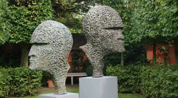 Zwei moderne Gartenskulpturen in Form von Köpfen aus Stein, im Grünen auf Podesten platziert.