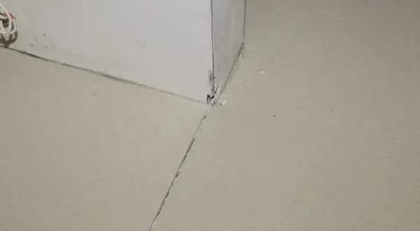Uszkodzona dylatacja posadzki w rogu pomieszczenia, widoczne pęknięcia i ubytki materiału.