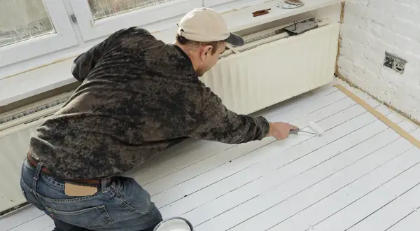 Un homme peint un plancher en bois blanc. Il utilise un rouleau et un pot de peinture. La question "peut on peindre du stratifié" est pertinente pour ce type de projet.