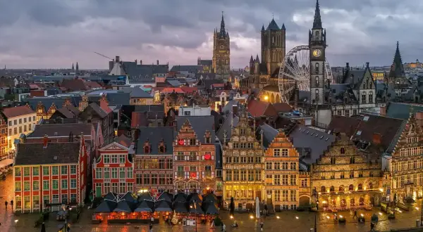 Gryning över Gent, Belgien. Historiska byggnader längs en kanal, med ett pariserhjul och klocktorn som reser sig mot en molnig himmel.