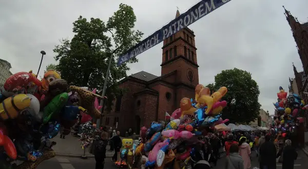 Kolorowe balony z postaciami z bajek i zwierzątkami, idealne gniezno atrakcje dla dzieci, na tle ceglanej wieży kościelnej i tłumu ludzi.