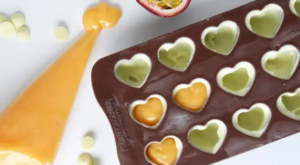 Gör egna enkla chokladpraliner recept med passionsfrukt och vit choklad i hjärtformad form. En påse med orange fyllning ligger bredvid.