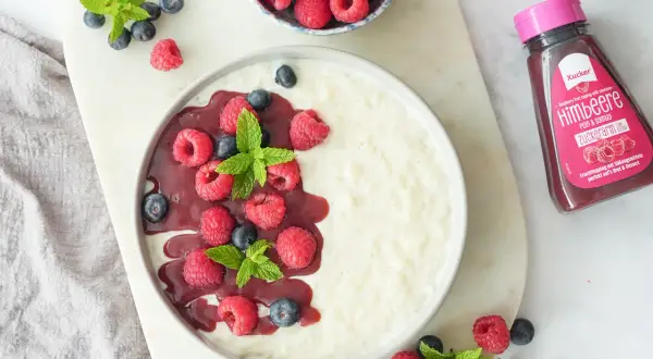 Cremiger Milchreis ohne Zucker, garniert mit frischen Himbeeren, Blaubeeren und Minze. Dazu eine Flasche zuckerarmer Himbeer-Fruchttoppping.