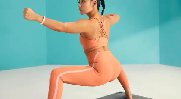 Mujer en ropa deportiva naranja practicando asanas de pie, con un brazo extendido y el otro doblado.