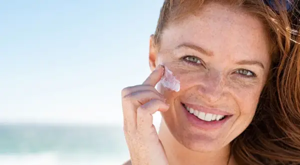 Frau mit Sommersprossen trägt Sonnenfluid auf ihr Gesicht auf, um ihre Haut vor der Sonne zu schützen.