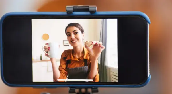 Frau hält Tasse und Keks, während sie ihr **iPhone als Webcam** für ein Kochvideo nutzt.