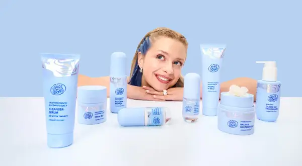 Uśmiechnięta dziewczyna z niebieskimi pasemkami otoczona kosmetykami Face Boom. Wśród nich kremy i sera, które dzięki caprylyl glycol odświeżają skórę.